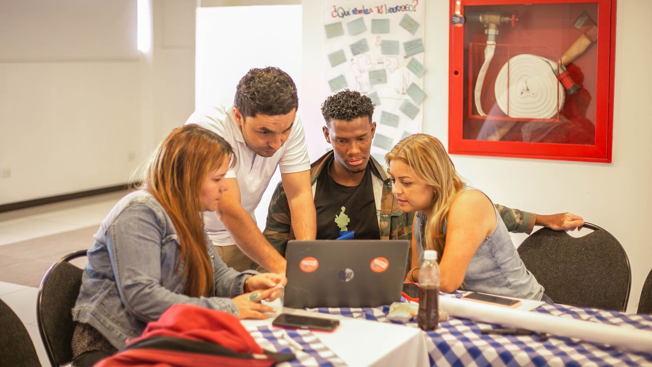 A diverse group of adults collaborating around a laptop during a team meeting in a professional setting.